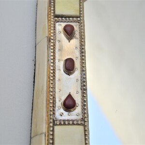 May include: A close-up of a decorative mirror frame with a silver background and three red gemstone accents. The frame is made of bone and has a gold-colored border.