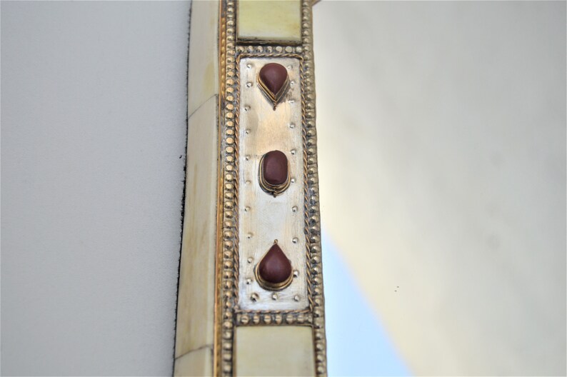 May include: A close-up of a decorative mirror frame with a silver background and three red gemstone accents. The frame is made of bone and has a gold-colored border.