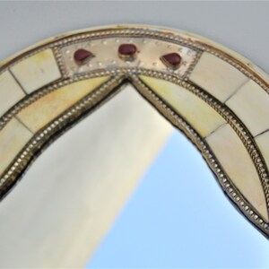 May include: A round, arched mirror with a decorative frame made of bone and brass. The frame is inlaid with small, red gemstones.