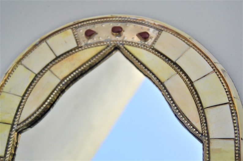May include: A round, arched mirror with a decorative frame made of bone and brass. The frame is inlaid with small, red gemstones.