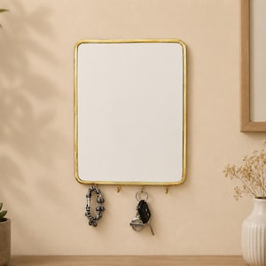 May include: A rectangular wall-mounted mirror featuring a thin, metallic gold-toned frame with rounded corners. The bottom edge of the frame includes four small, integrated hooks for hanging items such as a beaded bracelet and a set of car keys.