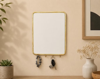 May include: A rectangular wall-mounted mirror featuring a thin, metallic gold-toned frame with rounded corners. The bottom edge of the frame includes four small, integrated hooks for hanging items such as a beaded bracelet and a set of car keys.