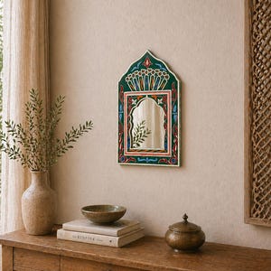 Pu&ograve; includere: Uno specchio da parete decorativo con una cornice dipinta di verde, rosso e blu. Lo specchio ha una parte superiore ad arco e una superficie rettangolare. &Egrave; appeso a una parete beige. Un tavolo di legno contiene un vaso, libri, una ciotola e un piccolo contenitore con coperchio.