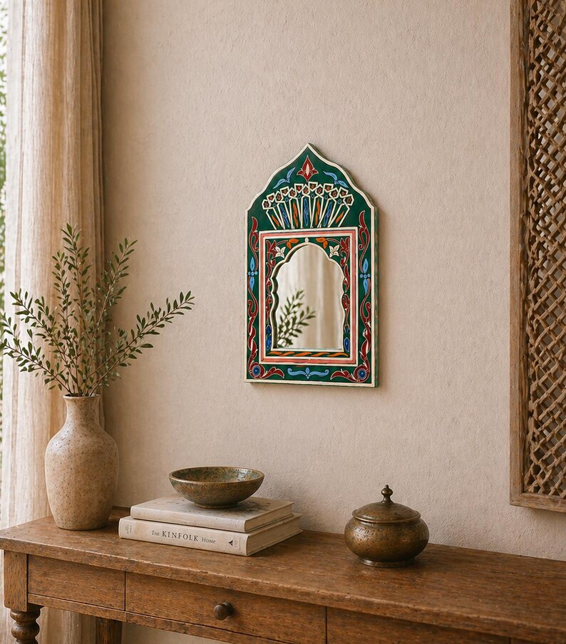 Pu&ograve; includere: Uno specchio da parete decorativo con una cornice dipinta di verde, rosso e blu. Lo specchio ha una parte superiore ad arco e una superficie rettangolare. &Egrave; appeso a una parete beige. Un tavolo di legno contiene un vaso, libri, una ciotola e un piccolo contenitore con coperchio.
