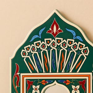 Pu&ograve; includere: Primo piano di una cornice decorativa ad arco per specchio. La cornice &egrave; dipinta in un verde intenso con motivi intricati in bianco, arancione, rosso e blu. Il design presenta elementi floreali e geometrici, creando un'estetica vibrante e ornata.
