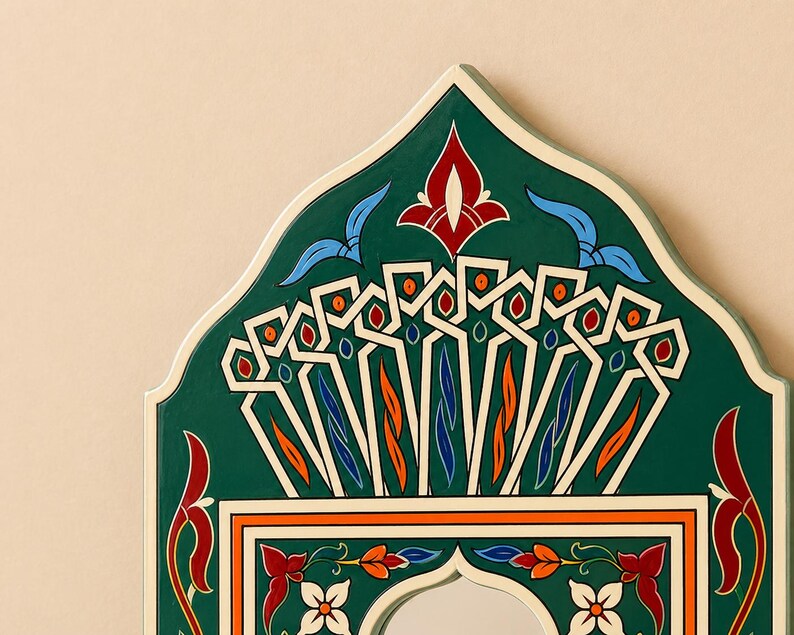 Pu&ograve; includere: Primo piano di una cornice decorativa ad arco per specchio. La cornice &egrave; dipinta in un verde intenso con motivi intricati in bianco, arancione, rosso e blu. Il design presenta elementi floreali e geometrici, creando un'estetica vibrante e ornata.