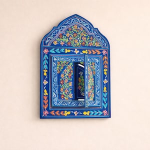Puede incluir: Un espejo de pared de madera pintado a mano de color azul vibrante con un diseño floral. El espejo tiene una parte superior arqueada y un marco rectangular con una pequeña puerta que se abre. El marco está adornado con flores y hojas coloridas.