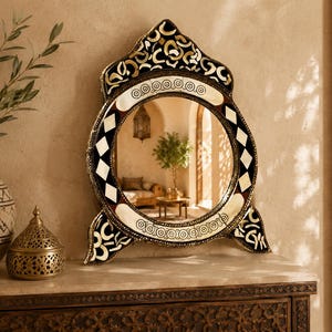May include: An ornate, decorative mirror with a circular reflective surface. The frame features a black, white, and gold geometric design. The mirror sits on a dark wooden surface, with a vase of greenery and a decorative metal container nearby.