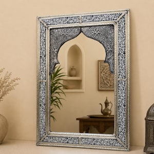 May include: A rectangular wall mirror featuring a silver-toned frame with intricate black and silver embossed scrollwork patterns. The upper portion of the mirror glass is framed by a decorative arch with detailed floral and geometric carvings.