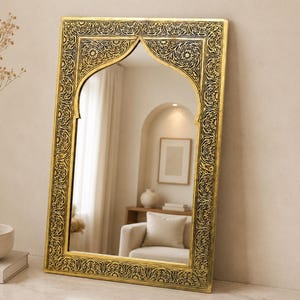 May include: A gold-framed mirror with an arched top and intricate floral detailing. The mirror reflects a room with a white sofa, artwork, and a vase with dried flowers. The mirror is leaning against a wall.