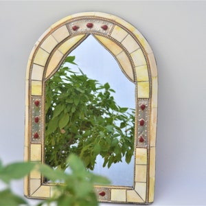 May include: A decorative arched mirror with a white and gold mosaic frame. The frame is accented with red jewel-toned accents. The mirror reflects a green leafy plant.