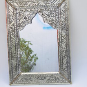 May include: A silver-colored metal mirror with an ornate, intricately carved frame. The frame features a traditional Moroccan design with a pointed arch at the top.
