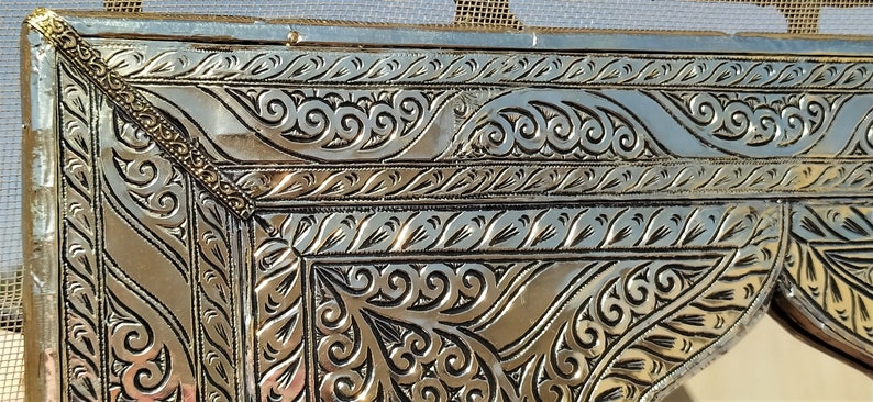 May include: Close-up of a silver metal frame with intricate embossed floral patterns. The frame has a gold border with a decorative design.