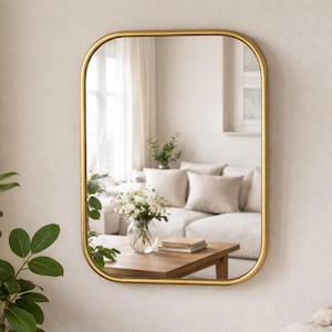 Pode incluir: Um espelho retangular com moldura dourada e cantos arredondados está pendurado numa parede de cor neutra. O espelho reflete uma cena de sala de estar com um sofá branco, almofadas e uma mesa de centro de madeira. Uma planta verde é visível à esquerda.