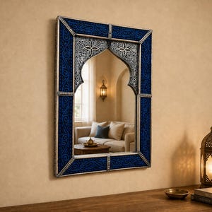 Puede incluir: Un espejo de pared rectangular con una parte superior arqueada, con marco plateado y borde azul oscuro estampado. El espejo refleja una habitación con un sofá y una lámpara decorativa.