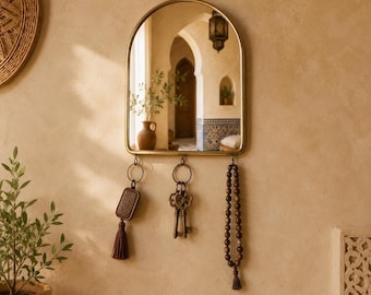 May include: A gold-framed arched mirror with a key holder. The mirror reflects an interior with arches and a hanging lantern. Below the mirror hang a key fob with a tassel, a set of keys, and a beaded necklace.