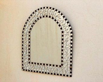 Op de afbeelding: Gewelfde mozaïekspiegel met een decoratieve rand van kleine, reflecterende, driehoekige tegels. De rand wisselt af tussen zwarte en witte vierkanten. De spiegel is aan een lichtgekleurde muur bevestigd.