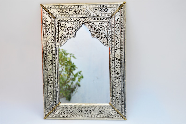 May include: A silver-colored metal mirror with an ornate, arched frame. The frame is decorated with intricate floral patterns and has a brass-colored trim.