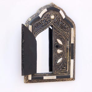 May include: Ornate, arched wall mirror with a hinged door. The mirror frame is black and gold with white and black accents. The door is open, revealing the mirror. The frame features intricate carvings and a decorative border.