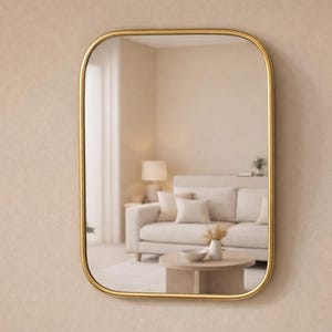 May include: A rectangular mirror with rounded corners and a gold frame hangs on a beige wall. The mirror reflects a living room scene with a sofa, coffee table, and lamp. The overall aesthetic is modern and minimalist.