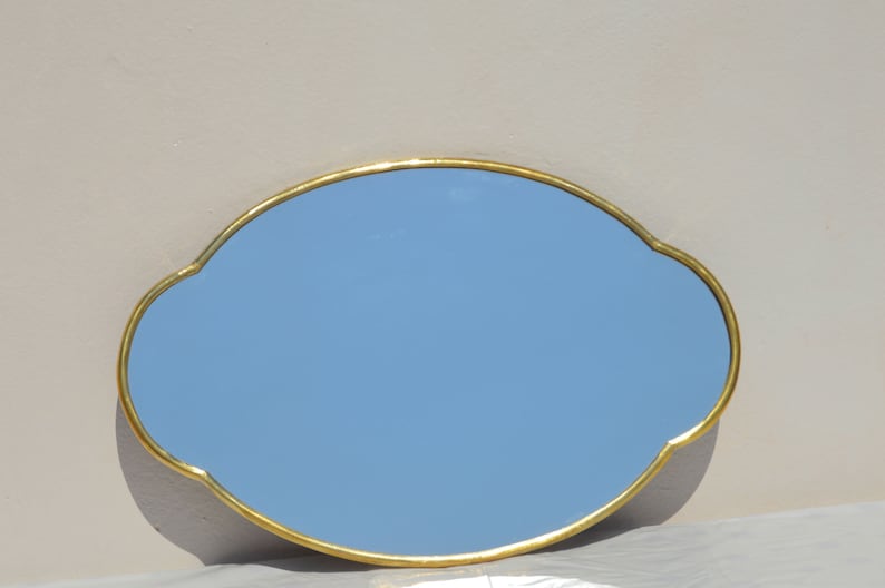 May include: A gold-framed oval mirror with a light blue reflective surface. The mirror has a decorative, scalloped edge.