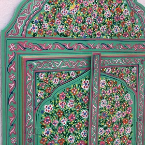 May include: A green wooden mirror with a decorative floral design painted in pink, white, and yellow. The mirror has a traditional Moroccan style.