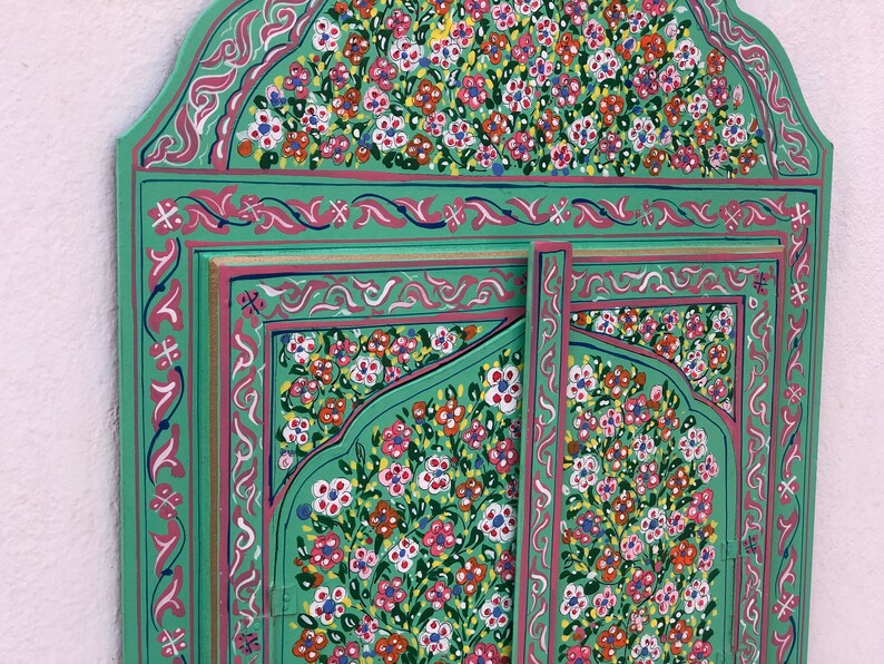 May include: A green wooden mirror with a decorative floral design painted in pink, white, and yellow. The mirror has a traditional Moroccan style.