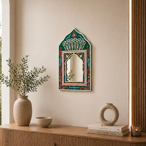 Pu&ograve; includere: Uno specchio da parete decorativo con una cornice dipinta di verde, rosso e blu. Lo specchio ha una parte superiore ad arco e una forma rettangolare. La cornice presenta intricati motivi floreali e geometrici. Lo specchio &egrave; appeso a una parete beige.