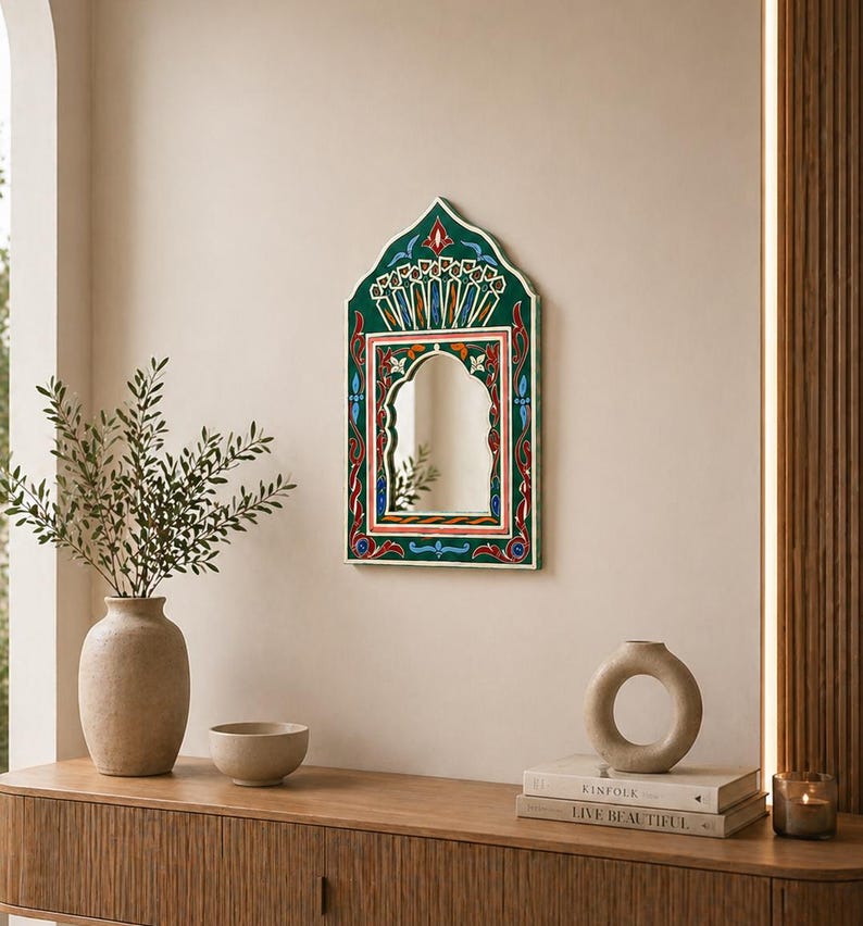 Pu&ograve; includere: Uno specchio da parete decorativo con una cornice dipinta di verde, rosso e blu. Lo specchio ha una parte superiore ad arco e una forma rettangolare. La cornice presenta intricati motivi floreali e geometrici. Lo specchio &egrave; appeso a una parete beige.