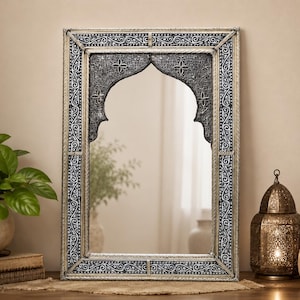 May include: A rectangular wall mirror featuring a silver-toned frame with intricate black and white scrollwork patterns. The mirror glass is set within a decorative arch-shaped insert that displays detailed floral carvings in a dark finish.