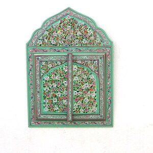 May include: A green wooden framed mirror with intricate floral designs painted in pink, white, and yellow. The mirror is divided into two sections with a decorative arch at the top.