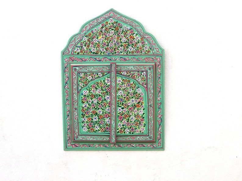 May include: A green wooden framed mirror with intricate floral designs painted in pink, white, and yellow. The mirror is divided into two sections with a decorative arch at the top.