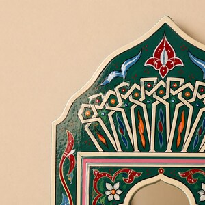 Pu&ograve; includere: Primo piano di una cornice di specchio decorativa ad arco. La cornice &egrave; dipinta in un verde vibrante con intricati motivi floreali e geometrici bianchi, rossi, arancioni e blu. I bordi sono delineati in bianco e rosa.