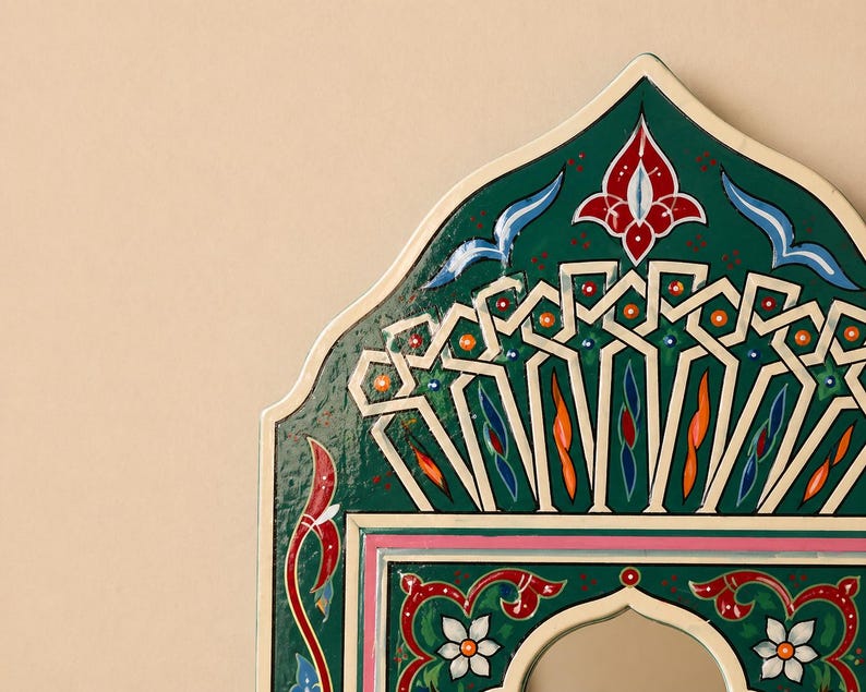 Pu&ograve; includere: Primo piano di una cornice di specchio decorativa ad arco. La cornice &egrave; dipinta in un verde vibrante con intricati motivi floreali e geometrici bianchi, rossi, arancioni e blu. I bordi sono delineati in bianco e rosa.