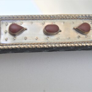 May include: Close-up of a silver-colored frame with three red teardrop-shaped stones. The frame has a decorative border and is made of metal.