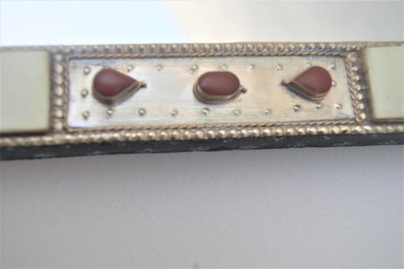 May include: Close-up of a silver-colored frame with three red teardrop-shaped stones. The frame has a decorative border and is made of metal.