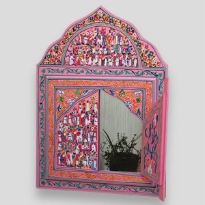 Puede incluir: Un espejo de madera pintado a mano de color rosa, con una parte superior arqueada y una puerta con bisagras. El marco y la puerta del espejo están adornados con motivos florales y figurativos coloridos. El espejo refleja una planta en una maceta. El diseño es ornamentado y decorativo.