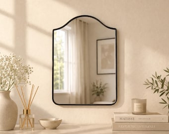 May include: A black-framed wall mirror with an arched top hangs on a beige wall. Below the mirror, a vase of white flowers, a reed diffuser, a small bowl, and two books are arranged. A lit candle sits on top of the books.