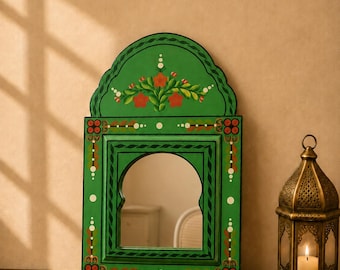 May include: A green wall mirror with an arched top and a central arched glass pane. The frame is decorated with hand-painted floral motifs in red, white, and brown, featuring symmetrical patterns along the sides and a floral spray at the top.