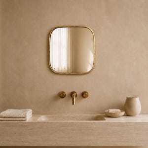 Op de afbeelding: Een vierkante spiegel met afgeronde hoeken en een dunne gouden lijst, gemonteerd boven een stenen wastafel. De wastafel is voorzien van een aan de muur gemonteerde gouden kraan met twee ronde knoppen, een gevouwen beige handdoek, een zeepbakje en een beige beker.