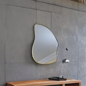 May include: A gold-framed, freeform mirror hangs on a gray wall. The mirror has an irregular, organic shape, resembling a cloud or a leaf.