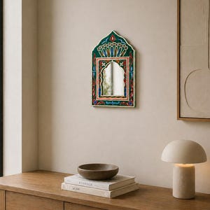Pu&ograve; includere: Uno specchio da parete decorativo con una cornice ornata verde, rossa e blu. Lo specchio ha una parte superiore ad arco e una forma rettangolare. Lo specchio &egrave; appeso a una parete beige. Un tavolo di legno con libri e una ciotola &egrave; in primo piano.
