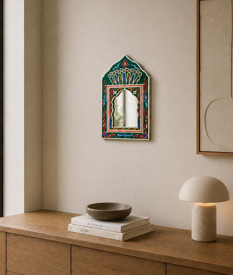 Pu&ograve; includere: Uno specchio da parete decorativo con una cornice ornata verde, rossa e blu. Lo specchio ha una parte superiore ad arco e una forma rettangolare. Lo specchio &egrave; appeso a una parete beige. Un tavolo di legno con libri e una ciotola &egrave; in primo piano.