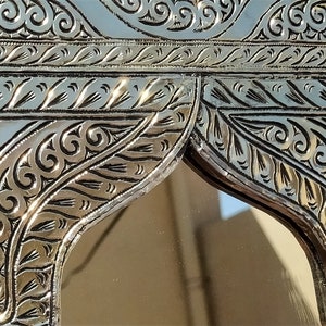 May include: A silver-colored metal mirror with an ornate, intricate design. The mirror has a rectangular frame with a decorative arch at the top. The design features swirling patterns and leaves.