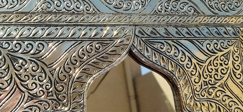 May include: A silver-colored metal mirror with an ornate, intricate design. The mirror has a rectangular frame with a decorative arch at the top. The design features swirling patterns and leaves.