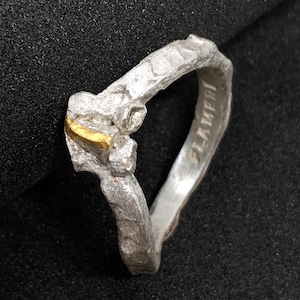 Könnte beinhalten: Ein Silberring mit einer texturierten, rauen Oberfläche und unregelmäßiger Form. Der Ring weist an der Oberseite einen kleinen, rechteckigen goldfarbenen Akzent auf. Die Innenseite des Rings ist mit dem Text FLAMERI in Großbuchstaben graviert.