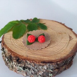 Pode incluir: Brincos de botão de framboesa vermelha com folhas verdes, apresentados em um pequeno cartão marrom com bordas onduladas. Os brincos são colocados em uma fatia de madeira rústica, com folhas verdes ao fundo.