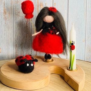 May include: A whimsical felted ladybug-themed display. It features a felt doll in a red dress holding a red balloon, a felt ladybug, and a candle decorated with a ladybug design. The items are arranged on a wooden base.