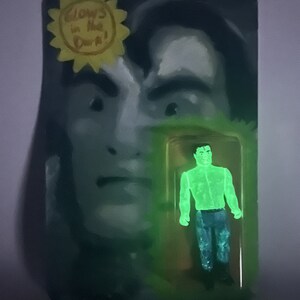 May include: A glow-in-the-dark action figure in a clear plastic package. The figure is a green humanoid with blue pants. The packaging features a painted face and the text "Glows in the Dark!" in a yellow sunburst.