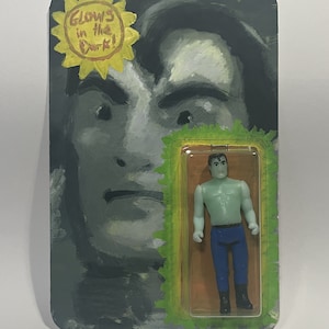 May include: A vintage-style action figure in a clear plastic case, mounted on a painted card. The card features a grayscale portrait and a yellow sunburst with the text "GLOWS in the Dark!". The figure is green with blue pants and black boots.